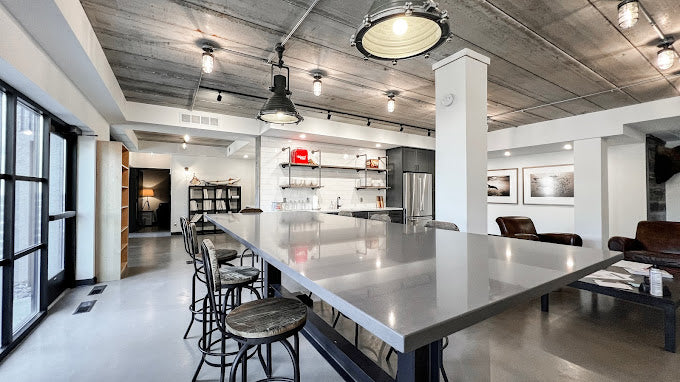 A row of MillerLights 'The Beacon' industrial caged ceiling lights integrated into an exposed electrical conduit network across a modern raw concrete ceiling in an open-concept office building.