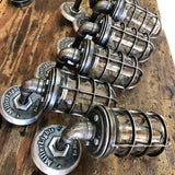 A row of handcrafted 'The Ward' industrial steel cage wall sconces lined up on a wooden workbench inside the MillerLights workshop, highlighting custom small-batch production and authentic iron pipe craftsmanship.