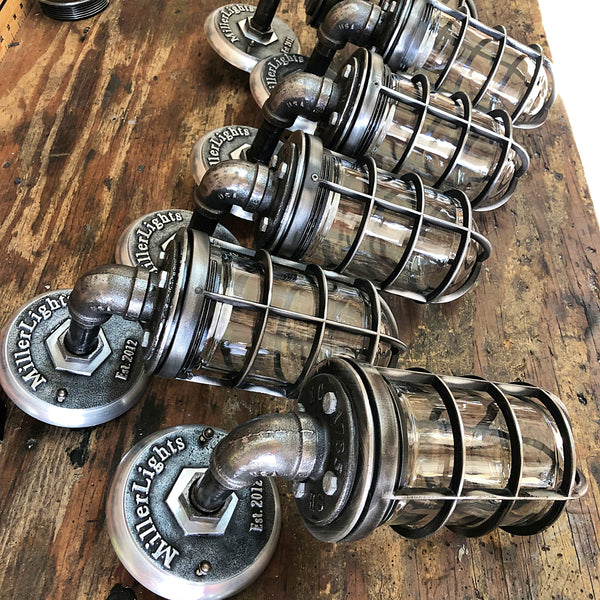 A row of handcrafted 'The Ward' industrial steel cage wall sconces lined up on a wooden workbench inside the MillerLights workshop, highlighting custom small-batch production and authentic iron pipe craftsmanship.