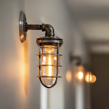 MillerLights 'The Ward' industrial wall sconce mounted in a long hallway, illuminated with a warm ambient glow from a clear glass jelly jar guard and an elongated vintage Edison bulb.