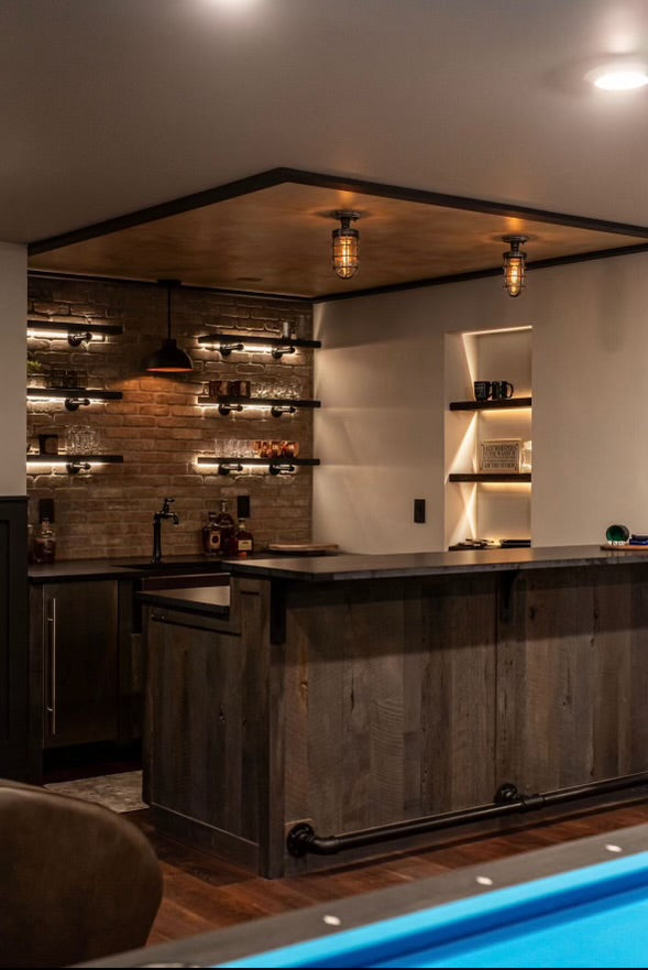 A pair of MillerLights 'The Beacon' industrial caged semi flush mount ceiling lights illuminating a rustic wood basement home bar counter with an exposed brick accent wall.