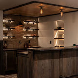 A pair of MillerLights 'The Beacon' industrial caged semi flush mount ceiling lights illuminating a rustic wood basement home bar counter with an exposed brick accent wall.