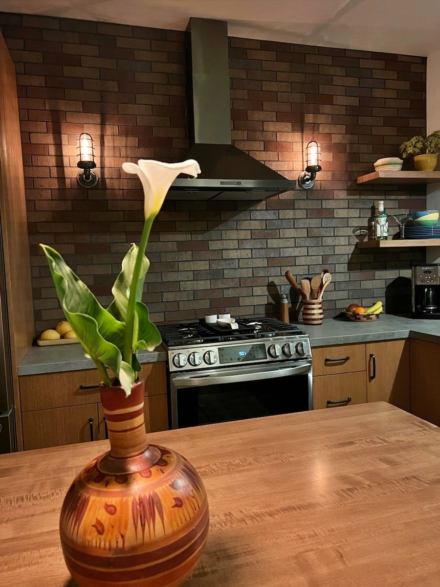 A pair of MillerLights 'The Ward' industrial single cage wall sconces installed facing upward on a dark brick accent wall, flanking a stainless steel kitchen range hood above a stove.