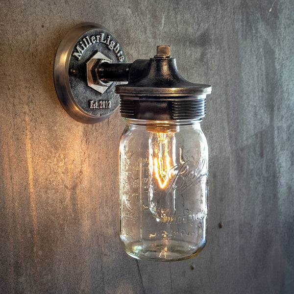 The mason jar light features a vintage-style design with a clear glass jar, The metal components have a weathered, black finish, complementing the textured, gray concrete wall background.