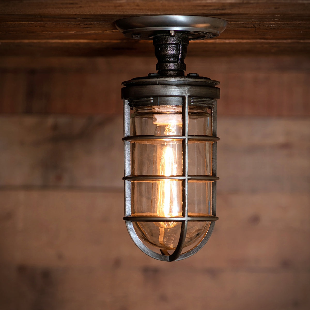 MillerLights 'The Beacon' industrial semi flush mount cage ceiling light fixture mounted flush onto a rustic wood barn beam, illuminated with a warm ambient glow from a clear glass jar and vintage filament bulb.
