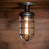 MillerLights 'The Beacon' industrial semi flush mount cage ceiling light fixture mounted flush onto a rustic wood barn beam, illuminated with a warm ambient glow from a clear glass jar and vintage filament bulb.