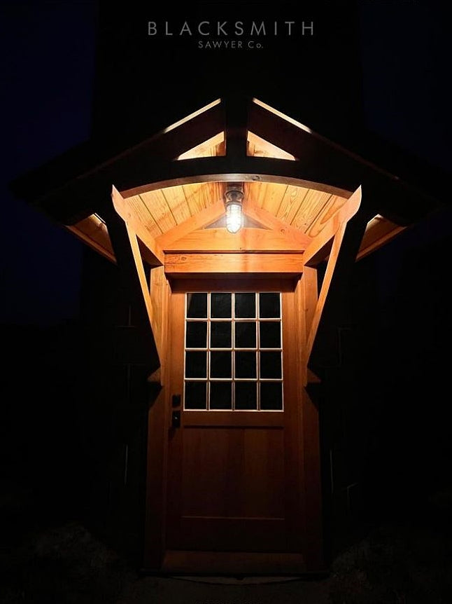 MillerLights 'The Beacon' industrial caged ceiling light fixture installed under a rustic wooden cabin porch portico, illuminating a wood and glass pane exterior front door at night.