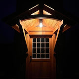 MillerLights 'The Beacon' industrial caged ceiling light fixture installed under a rustic wooden cabin porch portico, illuminating a wood and glass pane exterior front door at night.