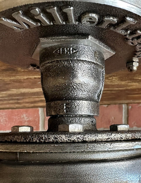 Close-up view of MillerLights 'The Beacon' industrial ceiling light mount, showcasing the cast iron canopy with stamped brand name, antiqued steel hex nuts, and heavy-duty iron pipe coupling joints.