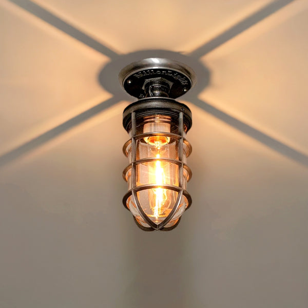 MillerLights 'The Beacon' model industrial semi flush mount ceiling light fixture, featuring an antiqued steel metal cage cover and clear glass jelly jar casting a dramatic warm ambient shadow pattern overhead.