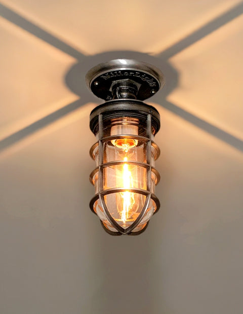 MillerLights 'The Beacon' model industrial semi flush mount ceiling light fixture, featuring an antiqued steel metal cage cover and clear glass jelly jar casting a dramatic warm ambient shadow pattern overhead.