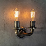 This fixture is a rustic wall-mounted light featuring two exposed Edison bulbs with warm, glowing filaments. The design incorporates a weathered, industrial-style metal pipe structure, with a central mounting plate engraved with 