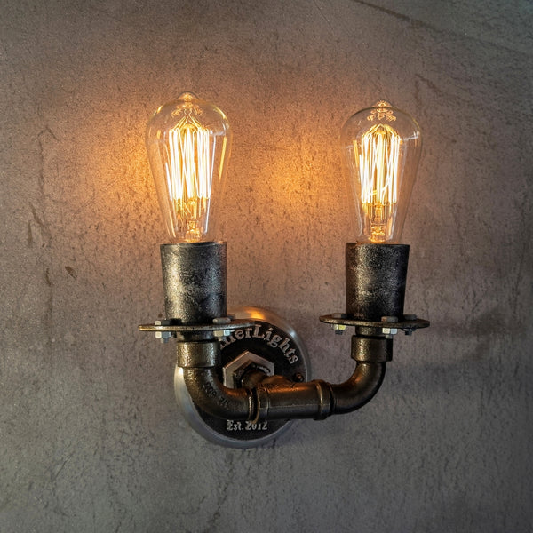 This fixture is a rustic wall-mounted light featuring two exposed Edison bulbs with warm, glowing filaments. The design incorporates a weathered, industrial-style metal pipe structure, with a central mounting plate engraved with "MillerLights Est. 2012." The bulbs are housed in dark, textured metal sockets, adding to the vintage aesthetic.