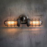 MillerLights industrial double cage wall sconce illuminated with a warm orange glow from vintage Edison bulbs, mounted on a textured gray concrete wall.