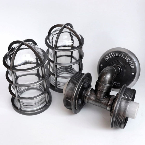 Disassembled view of the MillerLights industrial wall sconce showcasing the heavy-duty black iron pipe frame alongside two removable protective metal wire cages and clear glass globes.