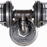 Close-up view of the MillerLights industrial vanity light fixture installed in an upside down position, showcasing the central iron pipe tee joint and the engraved brand wall plate.