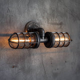 MillerLights iconic Joker model rustic industrial bathroom vanity lighting fixture, a handcrafted black iron pipe wall sconce with double protective wire cages over glowing Edison bulbs