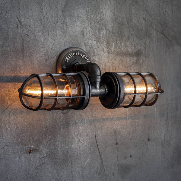 MillerLights iconic Joker model rustic industrial bathroom vanity lighting fixture, a handcrafted black iron pipe wall sconce with double protective wire cages over glowing Edison bulbs