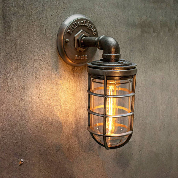 MillerLights 'The Ward' model industrial single cage wall sconce light fixture, featuring an antiqued darkened steel iron pipe arm and glass jelly jar guard illuminated against a textured concrete wall.